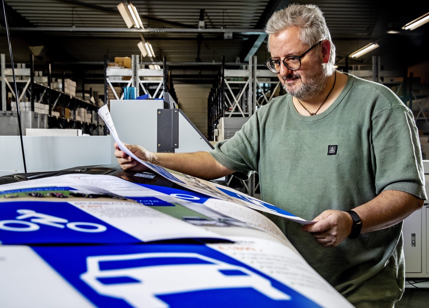 Een medewerker van Vogelaar houdt een vel drukwerk omhoog en controleert het, met de productiehal op de achtergrond.